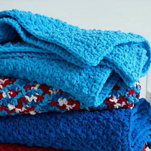 Une pile de trois couvertures au crochet, celle du haut en bleu clair, celle du milieu en rouge, blanc et bleu et celle du bas en bleu foncé.