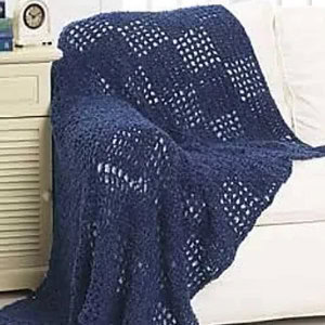 Une couverture afghane crochetée bleu foncé avec un motif carré est drapée sur un canapé blanc à côté d'une armoire de couleur claire avec une petite horloge sur le dessus.