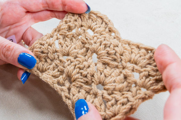 Gros plan de mains avec vernis à ongles bleu tenant un morceau de tissu crocheté beige, montrant son motif texturé et ouvert - parfait comme point au crochet pour couverture.