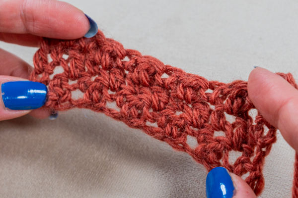 Gros plan de deux mains au vernis à ongles bleu tenant un morceau de tissu point au crochet pour couverture brun-rouge sur fond neutre.