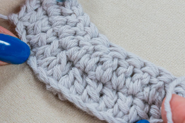 Gros plan de mains aux ongles peints en bleu tenant un petit morceau de tissu au crochet gris clair, montrant des points au crochet pour couverture détaillés sur un fond beige.