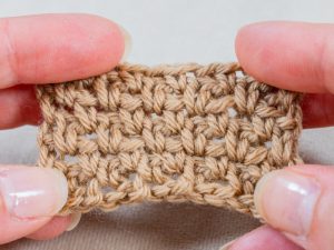 Gros plan des mains tenant un petit morceau de tissu au crochet de couleur beige, mettant clairement en évidence la texture et les points de riz au crochet sur un fond neutre.