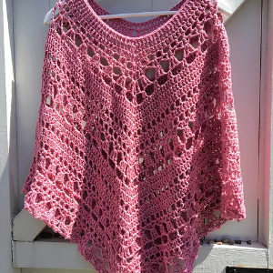 Un poncho rose crocheté à la main avec un motif ajouré en dentelle complexe est suspendu à un cintre blanc sur un fond en bois blanc.