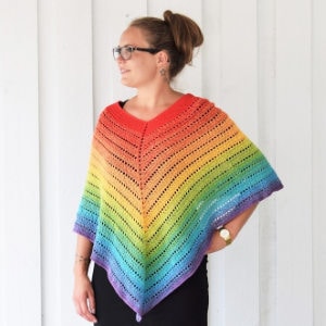 Une femme portant des lunettes et une robe noire se tient contre un mur blanc, modelant un poncho crocheté aux couleurs de l'arc-en-ciel avec un motif ajouré et un ourlet pointu.