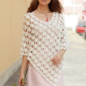 Une femme vêtue d'une robe rose clair et d'un poncho blanc en maille ajourée se tient dehors, sur une allée pavée, tenant une fleur d'une main. Son visage est partiellement hors cadre.
