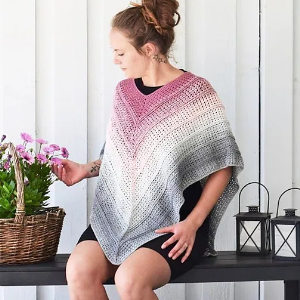 Une femme avec une coiffure en chignon porte un poncho au crochet dégradé en rose, blanc et gris, assise sur un banc à côté d'un panier de fleurs et de deux lanternes, devant un mur en bois blanc.