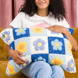 Une personne est assise sur un canapé jaune, tenant un coussin rectangulaire orné d'un motif floral crocheté bleu, blanc et jaune. Des rideaux roses apparaissent en arrière-plan.