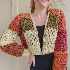 Une femme porte un cardigan patchwork coloré au crochet, orné de motifs carrés dans les tons orange, marron, vert, rouge et crème, sur un haut court noir et un pantalon noir. Elle sourit, la tête légèrement penchée.