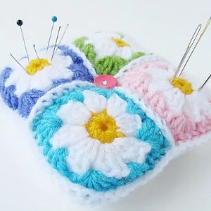 Un coussin à épingles coloré au crochet avec quatre carrés, chacun comportant une fleur blanche avec un centre jaune, contient plusieurs épingles et aiguilles à coudre.