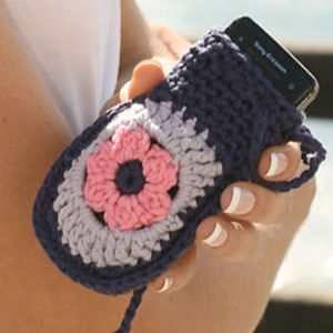 Une personne tient une pochette de téléphone crochetée ornée d'un motif floral bleu, gris et rose, recouvrant partiellement un téléphone Sony Ericsson noir. Elle porte une french manucure.