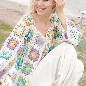 Une femme assise au bord de la plage, souriante, porte un pull coloré en crochet à carreaux de grand-mère sur un pantalon blanc. Le pull arbore des tons pastel de vert, bleu, violet, orange et crème.