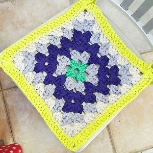 Housse de coussin carrée au crochet avec un centre vert, entouré de bleu foncé, de gris clair, de crème et d'une bordure jaune vif, posée sur un sol carrelé.