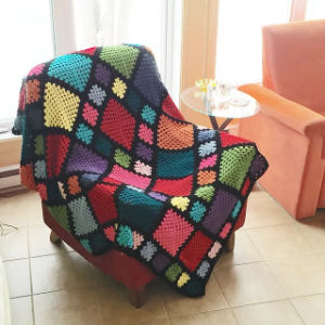 Une couverture patchwork colorée au crochet avec différents carrés drapée sur un fauteuil rouge dans une pièce lumineuse et ensoleillée avec du carrelage et une chaise orange à proximité.
