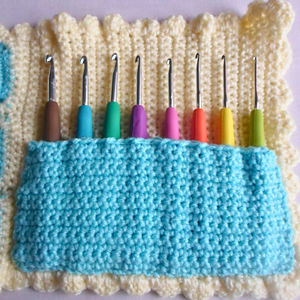 Un ensemble de huit crochets avec des poignées colorées rangés dans un support crocheté bleu clair, placé sur un tapis crocheté jaune avec un bord festonné.