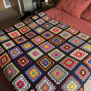 Un lit recouvert d'une couverture au crochet faite à la main avec des carrés de grand-mère colorés bordés de noir, étalés sur une courtepointe couleur rouille dans une chambre confortable.