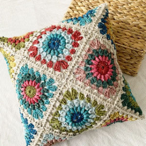 Coussin carré au crochet aux motifs floraux colorés en bleu, rose, vert et rouge. Ce coussin texturé, réalisé à la main, est posé sur un tissu léger, à côté d'une surface tissée.