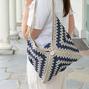 Une personne portant une chemise blanche porte un grand sac au crochet fait main à carreaux bleu marine et beige avec une fermeture à bouton sur son épaule.