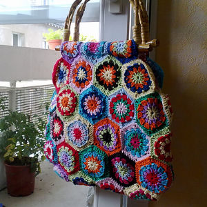 Un sac fourre-tout en crochet coloré, orné de motifs hexagonaux aux couleurs vives, est suspendu à une poignée de porte. Une plante en pot et la balustrade d'un balcon sont visibles en arrière-plan.