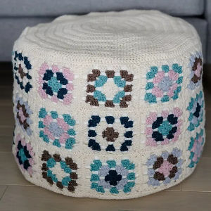 Un pouf rond crocheté avec une base blanche, décoré de motifs carrés de grand-mère multicolores dans des tons de bleu, rose, marron et gris, posé sur un plancher en bois.
