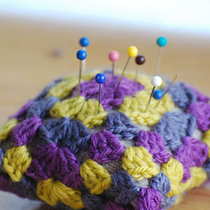 Gros plan d'un coussin à épingles tricoté en fil violet, jaune et gris, contenant plusieurs épingles à coudre à têtes rondes colorées.