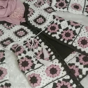 Un projet de crochet avec des carrés de grand-mère en fil rose, blanc et gris est disposé sur une surface, avec une écharpe assortie, une pelote de fil et une écharpe ou un tissu rose à proximité.