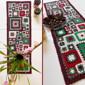 Un chemin de table au crochet coloré avec des motifs carrés rouges, verts, blancs et bleu sarcelle bordés de bordeaux, présenté sur une surface blanche avec une fleur rose et un bol de pot-pourri à proximité.