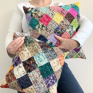 Une personne vêtue d'une chemise blanche à manches longues tient deux coussins crochetés en patchwork de petits carrés multicolores. Le fond est uni et clair.