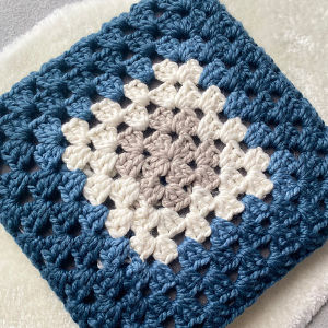 Un carré de grand-mère crocheté avec un centre gris clair, entouré de fil blanc, bleu clair et bleu foncé, présenté sur une surface douce et de couleur claire.
