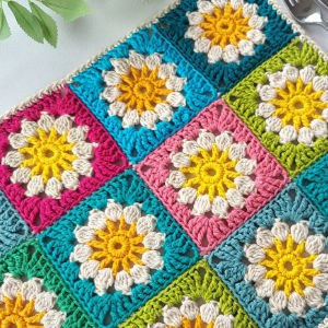 Carrés de grand-mère au crochet colorés présentant des motifs floraux en fil bleu, rose, vert et jaune, disposés dans une grille soignée sur une surface plane avec des feuilles vertes dans le coin.