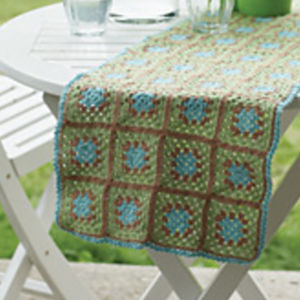 Un chemin de table au crochet avec des carrés de grand-mère verts, marron et bleus drapés sur une table d'extérieur ronde blanche avec des verres et un pichet sur le dessus.