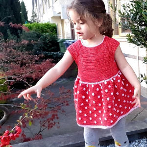 Une jeune fille vêtue d'une robe rouge à pois blancs tend la main vers des feuilles rouges sur un buisson à l'extérieur, debout sur un trottoir près de la verdure et des bâtiments.