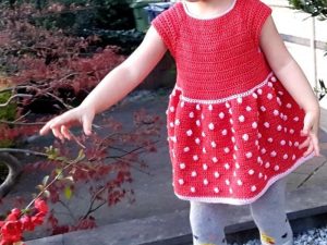 Un jeune enfant vêtu d'une robe rouge bébé au crochet à pois blancs tend la main vers une plante à l'extérieur, debout sur une surface pavée avec de la verdure en arrière-plan.