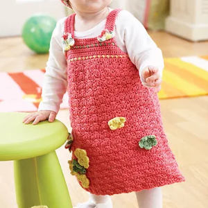 Une petite fille, vêtue d'une robe rose au crochet ornée d'appliqués de fleurs jaunes et vertes, se tient à l'intérieur, la main posée sur un tabouret vert clair. Elle porte un t-shirt blanc à manches longues sous sa robe.