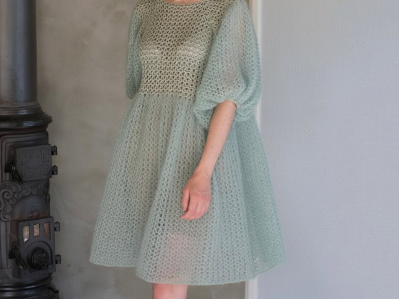 Une personne portant une robe en maille aérée vert clair avec des manches bouffantes, rappelant une robe au crochet, se tient à l'intérieur à côté d'un poêle à bois noir vintage et d'un mur de couleur claire.