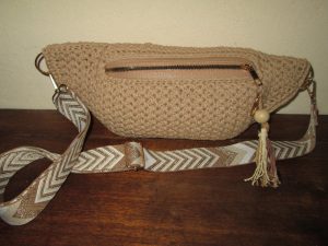 Sac banane beige au crochet avec une fermeture éclair, un pompon décoratif et une large bandoulière réglable à motifs, reposant sur une surface en bois contre un mur de couleur claire.