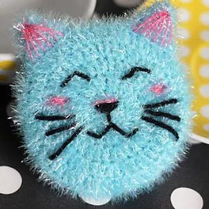 Une brosse à récurer bleue et duveteuse en forme de chat avec des moustaches, des yeux et un nez noirs brodés, ainsi que des détails roses sur les oreilles et les joues, repose sur une surface à pois noirs et blancs avec un objet jaune en arrière-plan.