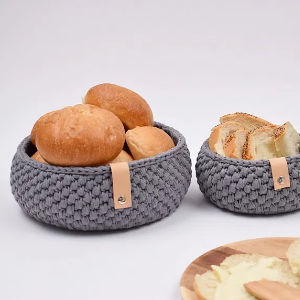 Deux paniers gris en tricot, ornés d'étiquettes en cuir beige, contiennent des petits pains et du pain de mie. Une planche de bois garnie de fromage ou de pâte à tartiner est partiellement visible au premier plan. L'arrière-plan est blanc uni.