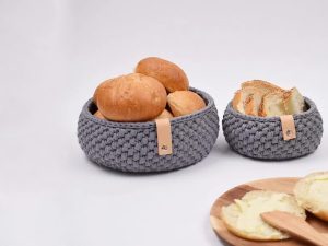 Deux paniers en tricot gris, inspirés du tutoriel de corbeille au crochet, contiennent des petits pains frais et du pain de mie. Au premier plan, une assiette en bois présente des tranches de pain beurrées et un petit tartineur en bois, le tout sur un fond blanc uni.