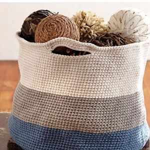 Un panier crocheté à rayures bleues, grises et blanches, rempli de boules décoratives en matériaux naturels, repose sur une surface en bois.