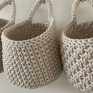 Trois paniers beiges, tissés à la main en fil tressé épais, sont suspendus côte à côte sur un fond clair. Chaque panier présente une forme arrondie et une poignée robuste au sommet.