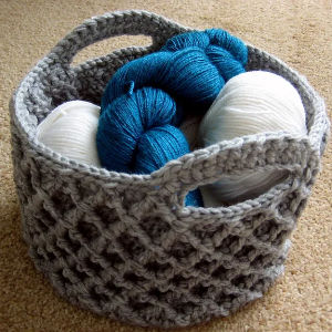 Un panier crocheté gris contient deux pelotes de fil blanc et une écheveau de fil bleu, posés sur un tapis beige.