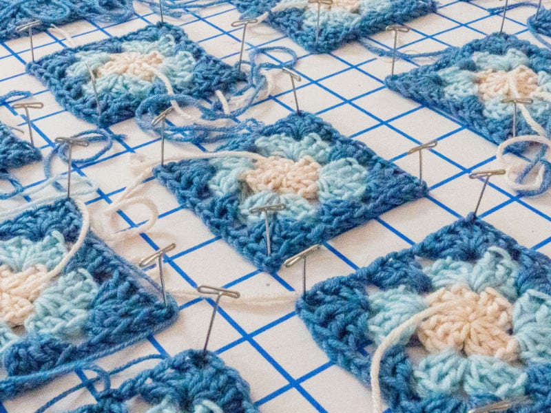 Des carrés de grand-mère crochetés dans des tons bleus et crème sont épinglés sur une planche à bloquer quadrillée, avec des queues de fils libres visibles. Les carrés, qui font partie d'un projet de tuto granny au crochet, sont en train d'être façonnés et préparés pour l'assemblage.