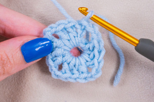 Gros plan sur une main au vernis à ongles bleu tenant un hexagone crocheté bleu clair, à côté d'un crochet doré au manche gris - parfait pour illustrer un tuto granny au crochet.