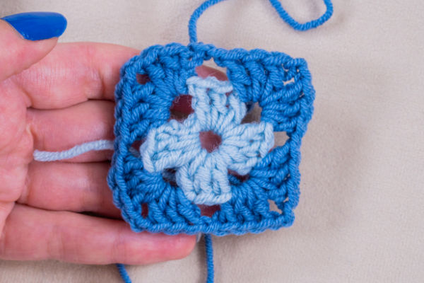 Une main avec du vernis à ongles bleu tient un carré de grand-mère crocheté, avec un motif de croix bleu clair au centre et une bordure bleu moyen, parfait pour suivre un tuto granny au crochet, avec des queues de fils visibles.