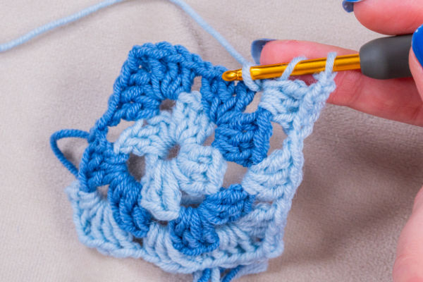 Gros plan d'une main tenant un crochet et travaillant avec du fil bleu et bleu clair pour créer un motif granny square sur un fond de tissu neutre - parfait pour un projet de crochet tuto granny au.