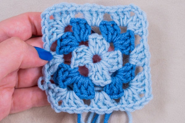 Une main avec du vernis à ongles bleu tient un carré au crochet réalisé avec des fils bleu clair et bleu moyen, présentant un motif géométrique sur un fond beige - une inspiration parfaite pour votre prochain tuto granny au crochet.