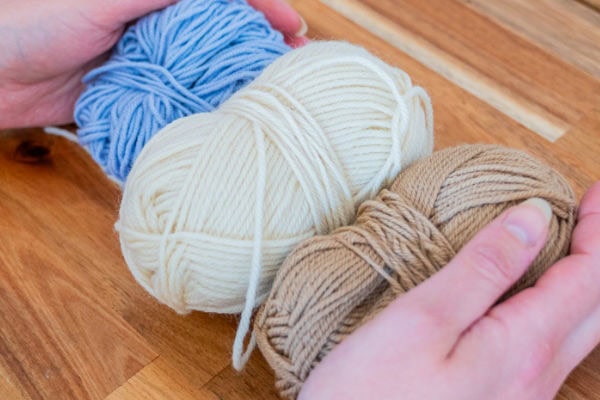 Deux mains tenant trois pelotes de laine en bleu, blanc et beige sur une surface en bois - parfait pour commencer votre prochain tuto granny au crochet.