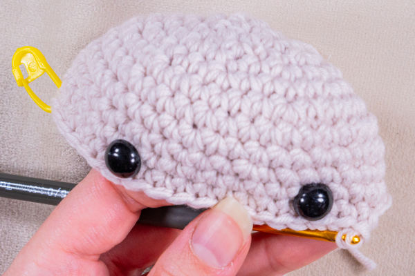Une main tient une peluche au crochet partiellement terminée avec un fil beige clair, deux yeux de sécurité noirs et un marqueur de point jaune attaché sur le côté. Un crochet est inséré dans les mailles du bas.