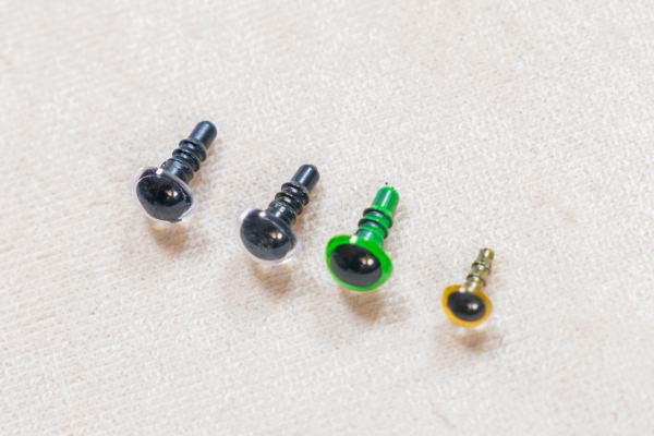 Quatre petites vis de tailles différentes sont alignées sur une surface en tissu léger. Idéales pour fixer des yeux de sécurité, les deux plus grandes vis sont argentées, la troisième est argentée avec un anneau vert et la plus petite est dorée avec un anneau jaune.