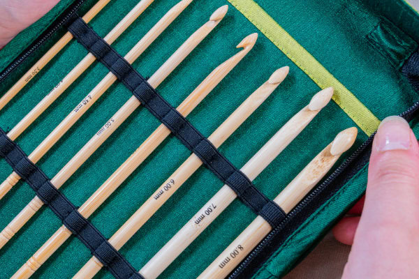 Un ensemble de crochets en bambou de différentes tailles est soigneusement rangé dans un étui en tissu vert, tenu ouvert par deux mains. Les crochets, qui ressemblent à ceux des collections Crochet Clover, sont fixés à l'aide d'élastiques noirs.